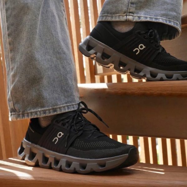 Person wearing black On running shoes with distinctive segmented sole stepping on wooden stairs, jeans cuffs visible.