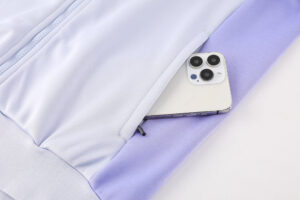 100095de | UAE SPORT STORE Light purple athletic jacket with a zip pocket holding a smartphone with a triple-camera back visible outside the pocket, resting on fabric.