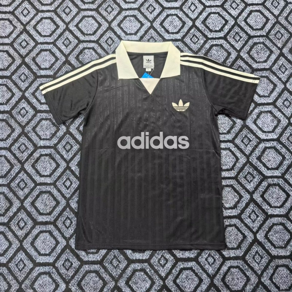 Black Adidas polo with white collar and three white stripes on sleeves, large Adidas logo and text on front resting on a patterned carpet.