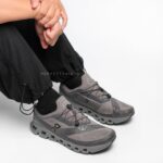 Gray athletic sneakers with chunky soles, laced, worn by a seated person in black pants against a white background.