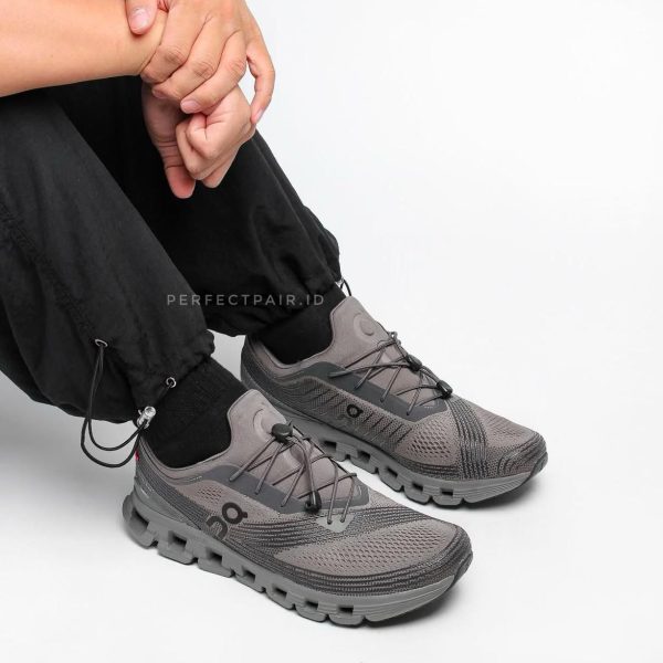 Gray athletic sneakers with chunky soles, laced, worn by a seated person in black pants against a white background.
