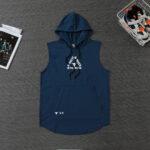 Navy blue sleeveless hoodie with drawstrings and front pocket laid on a dark wooden surface, with sneakers in a basket and a magazine nearby.