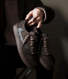 Brown suede sneakers with blue side panels are being held by a hand wearing a silver ring.
