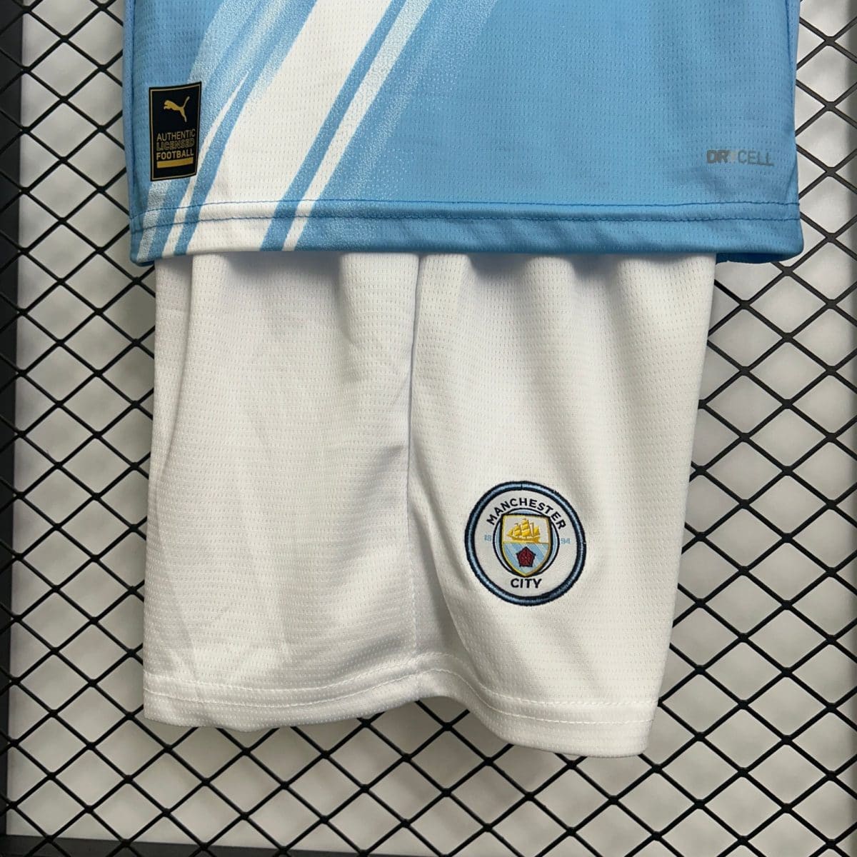Close-up of a light blue Manchester City football jersey sleeve with club crest and a diagonal white stripe on a chain-link background.