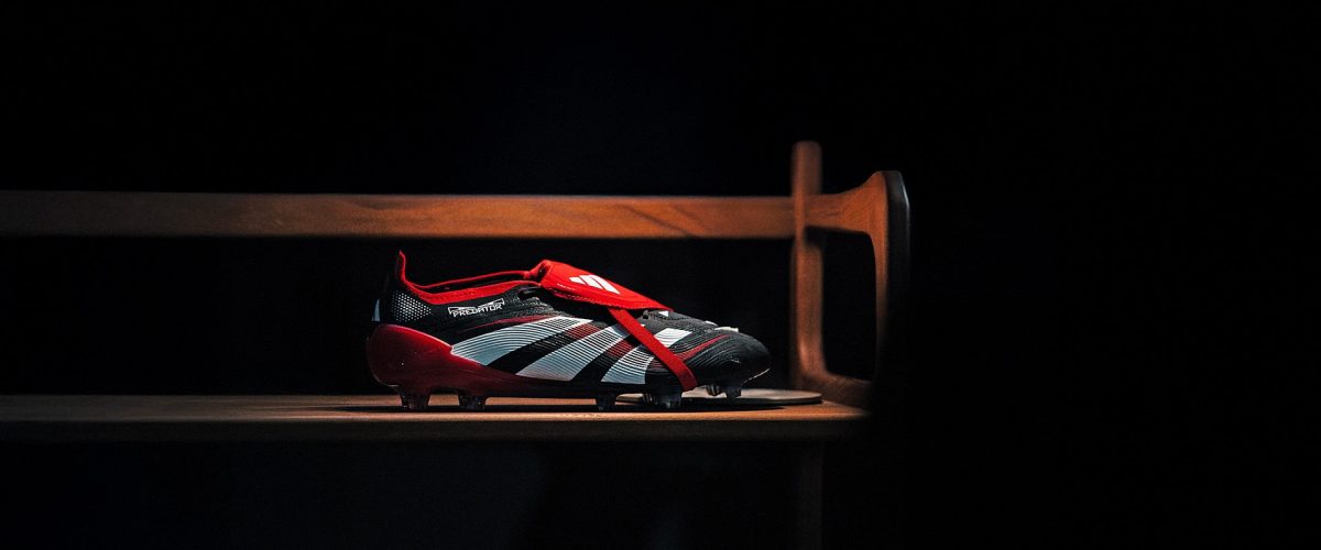 Red and black soccer cleat with white stripes labeled Predator, resting on a wooden bench in a dark room with dramatic lighting.