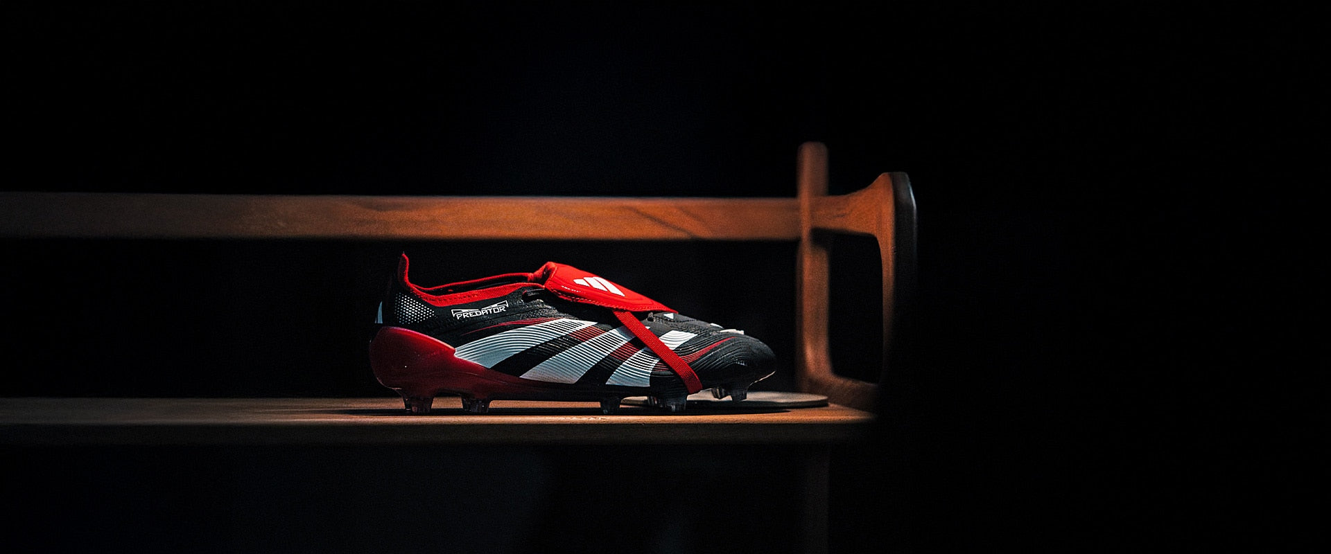 1026312_Creative_Main_1945798 | UAE SPORT STORE Red and black soccer cleat with white stripes labeled Predator, resting on a wooden bench in a dark room with dramatic lighting.