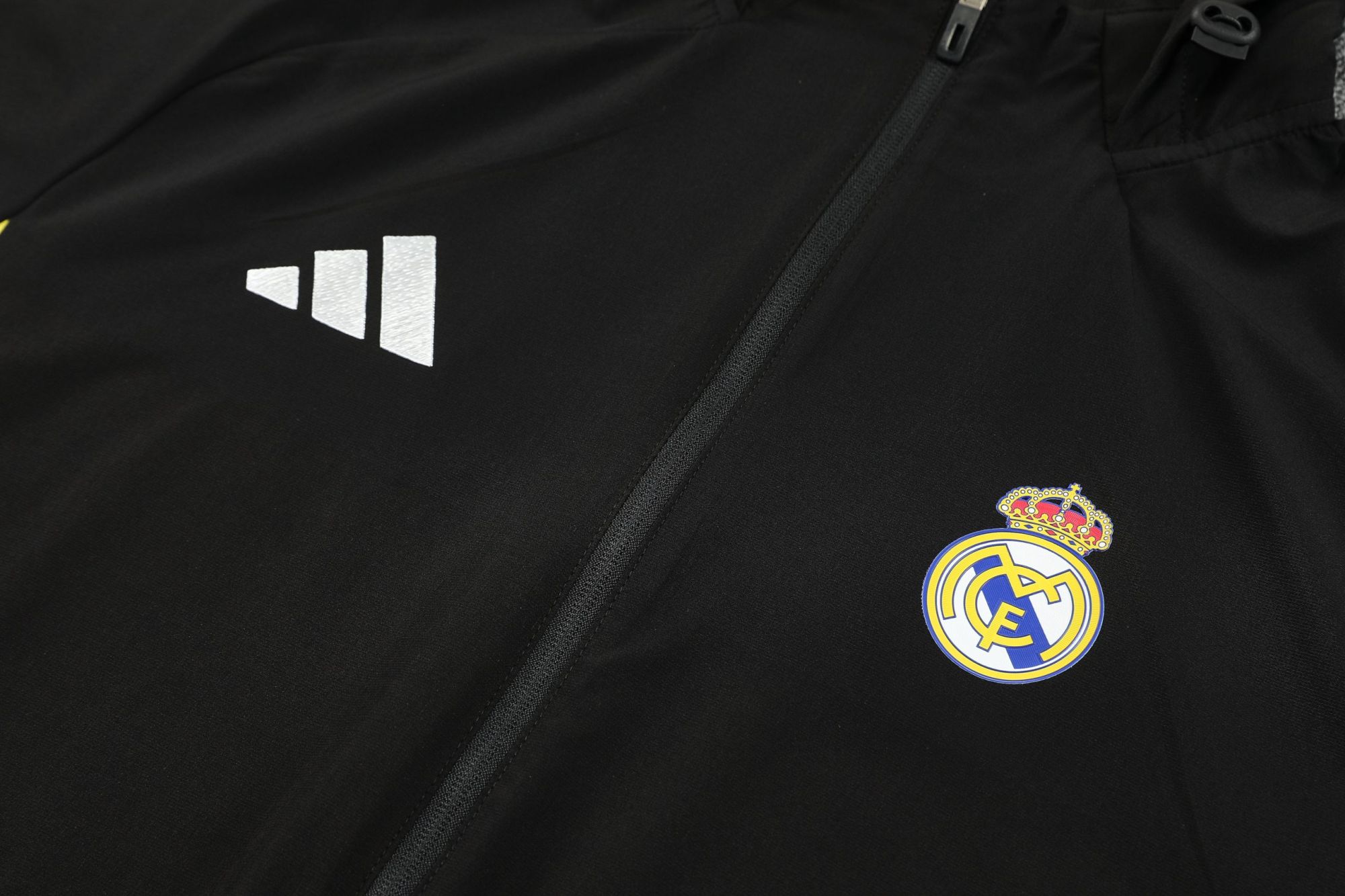 1029a2fe | UAE SPORT STORE Black athletic jacket close-up showing a white Adidas logo near a zipper and Real Madrid crest on the lower right.