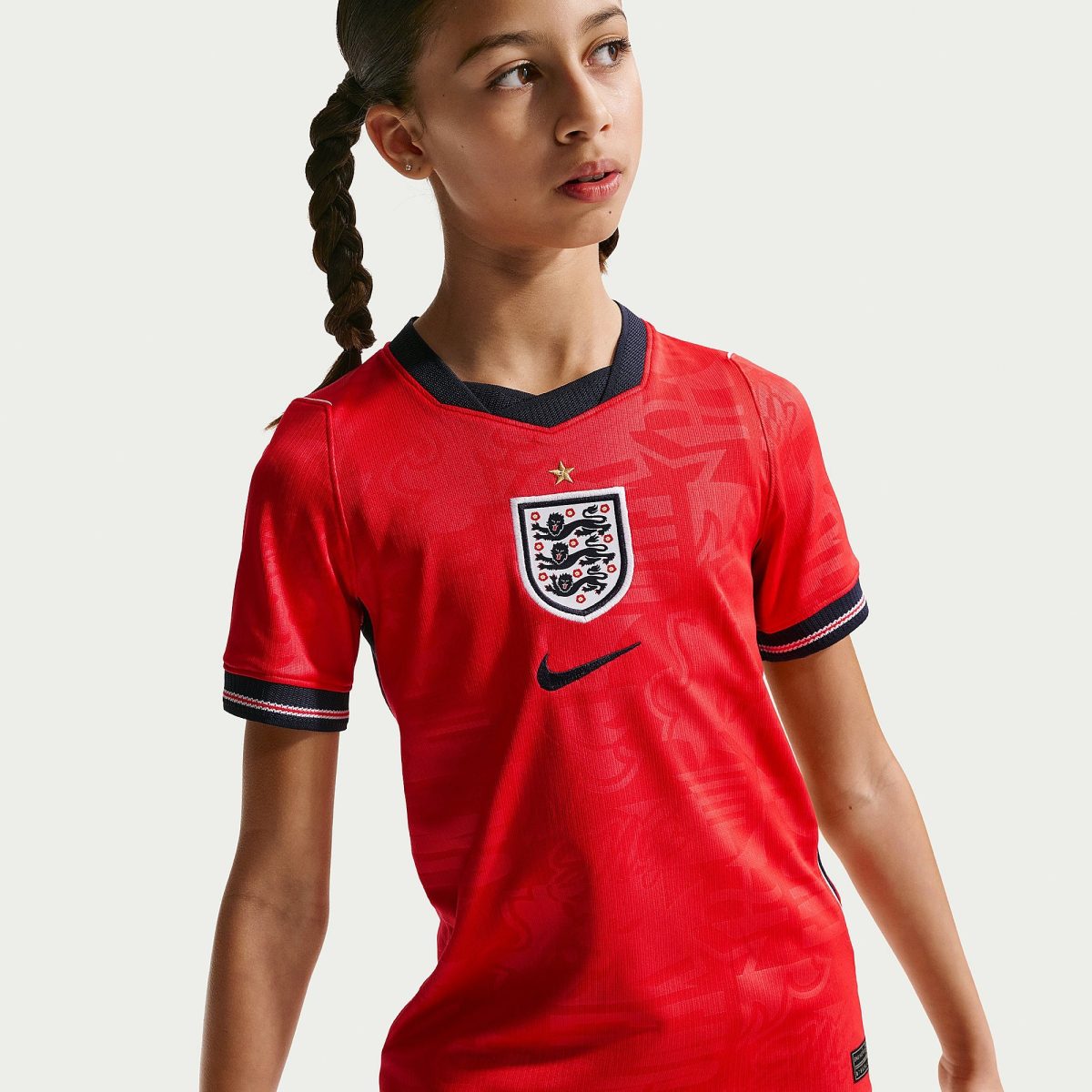 Young woman with braided hair wearing a red England football jersey with a crest, looking to the right.