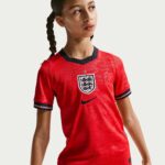 Young woman with braided hair wearing a red England football jersey with a crest, looking to the right.