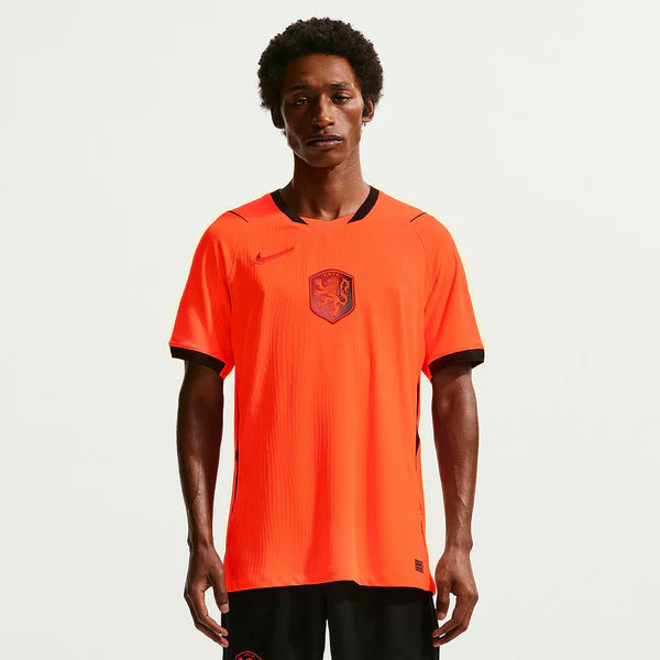 Male model wearing bright orange soccer jersey with black trim and crest, facing camera.