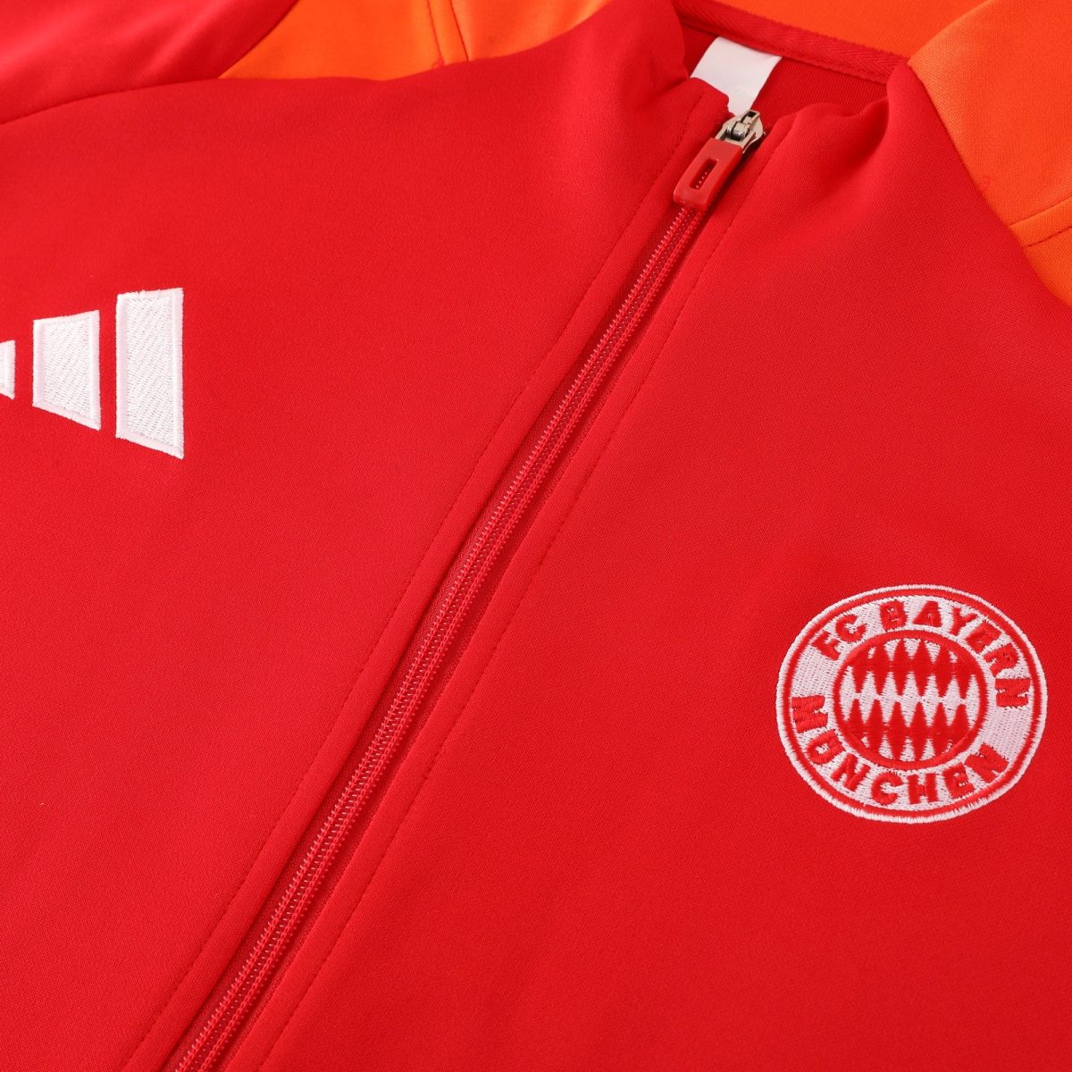 Close-up of a red sports jacket zipper with white stitched patches and a Bayern Munich crest visible in the bottom-right corner.