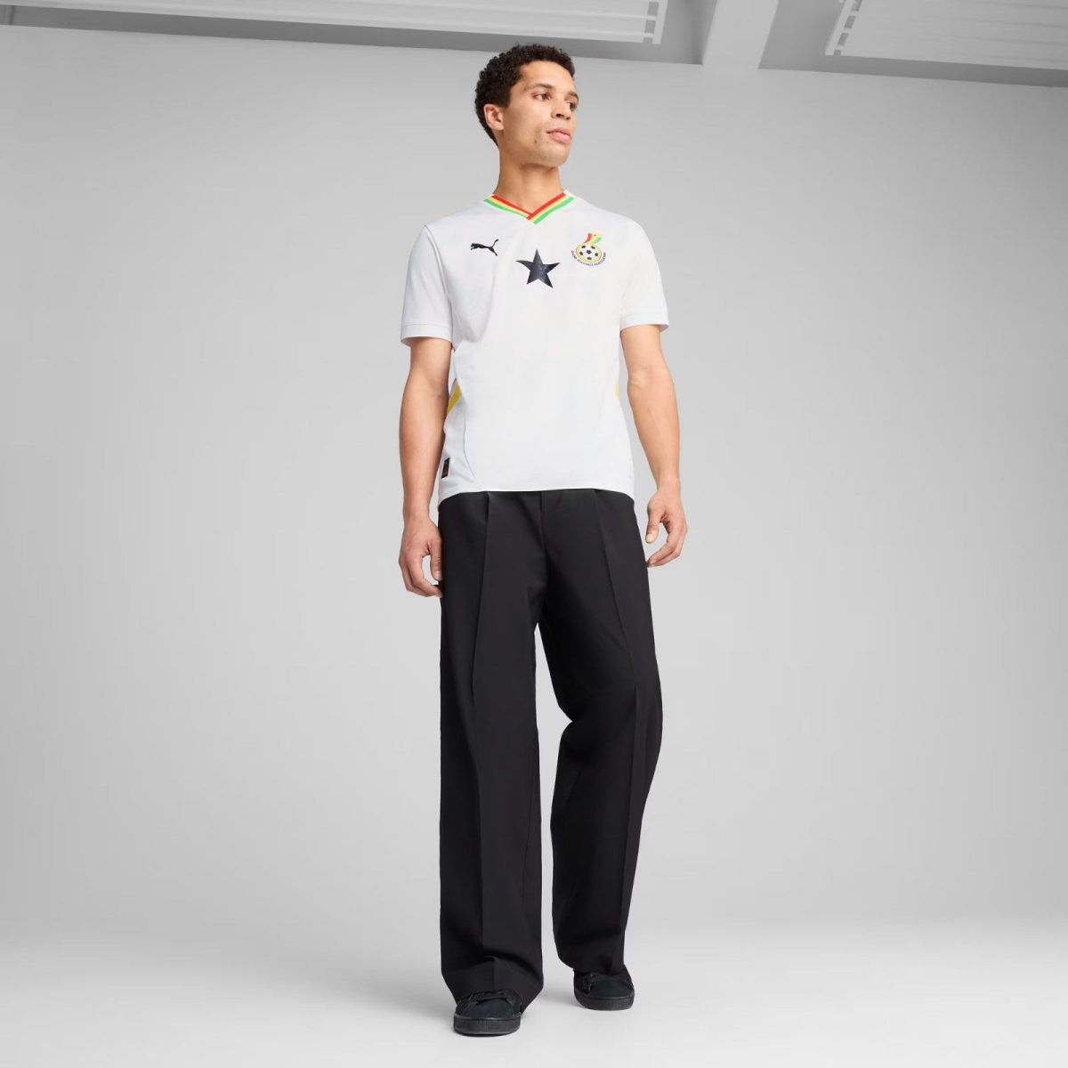 Male model wearing a white football jersey with Ghana colors at the collar, black star emblem, and team badge, paired with black pants in a studio.
