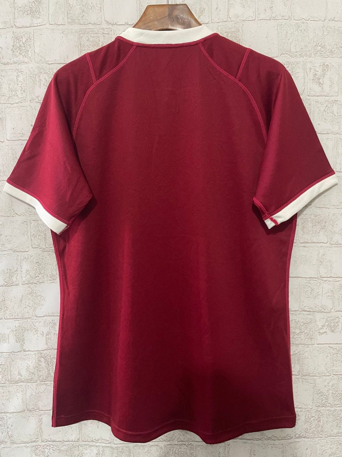 Back view of a burgundy polo shirt with a white collar and cuffs on a wooden hanger against a light brick wall.