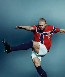 Soccer player in a red Norway jersey performing a high kick mid-air against a blue background.