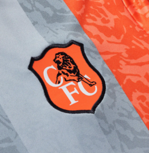 Close-up of an orange shield crest with a black lion and white letters 'FC' on gray fabric.