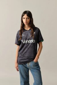 12_W_Home_Jersey_Venezia_3_f6d6c935-9d81-4ef4-a226-c91bdd289edf | UAE SPORT STORE عارضة ترتدي قميص فينيتسيا رياضيًا باللون الرمادي الداكن بنقشة رخاميّة، وتلتقط صورة أمام خلفية محايدة.