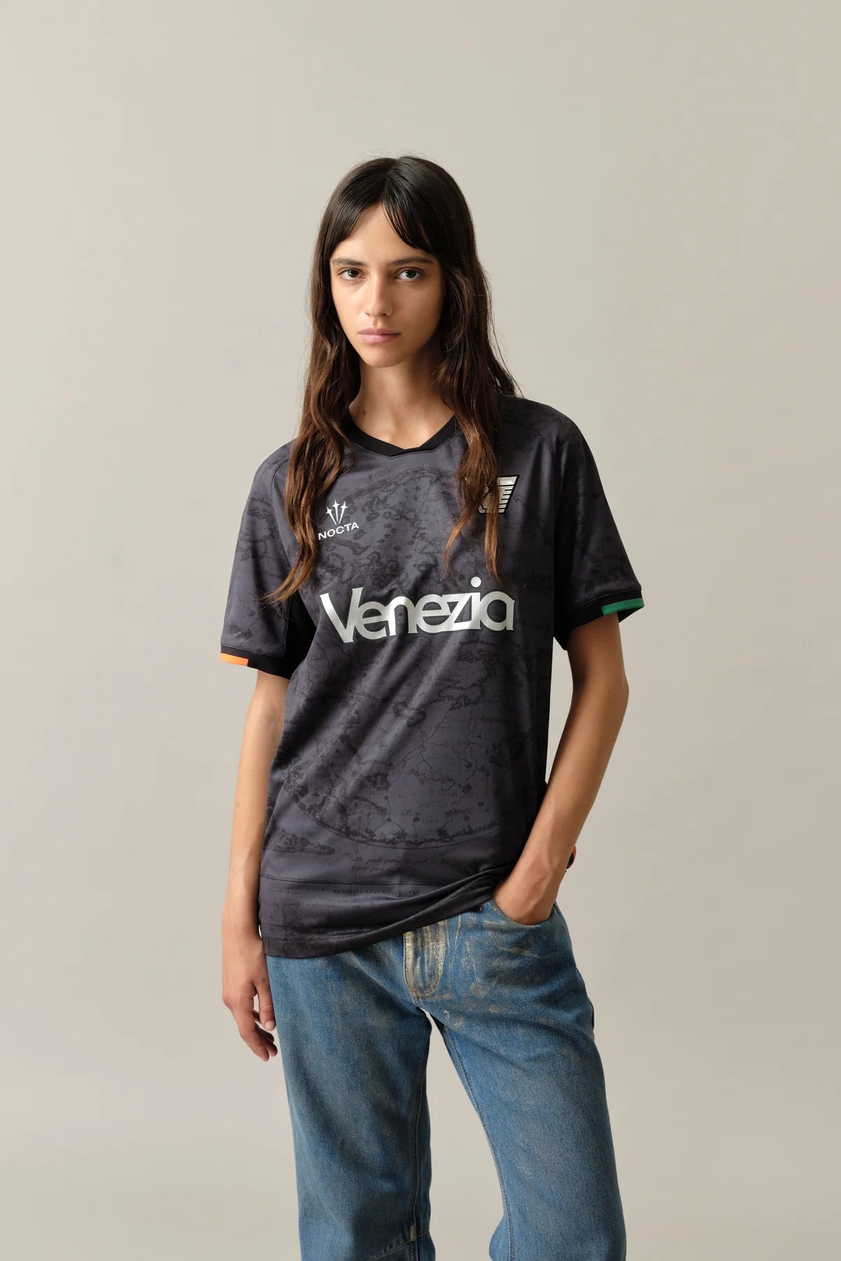 12_W_Home_Jersey_Venezia_3_f6d6c935-9d81-4ef4-a226-c91bdd289edf | UAE SPORT STORE عارضة ترتدي قميص فينيتسيا رياضيًا باللون الرمادي الداكن بنقشة رخاميّة، وتلتقط صورة أمام خلفية محايدة.