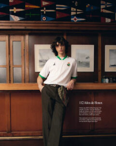 12_cweb_400x_c5528c40-5763-4731-93f3-a44f24bcfff2 | UAE SPORT STORE Young person with curly hair in a white polo with green trim leaning against a wooden bar beneath nautical flags.