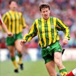 Soccer player in a green and yellow striped kit sprinting with the ball during a match on a crowded field.