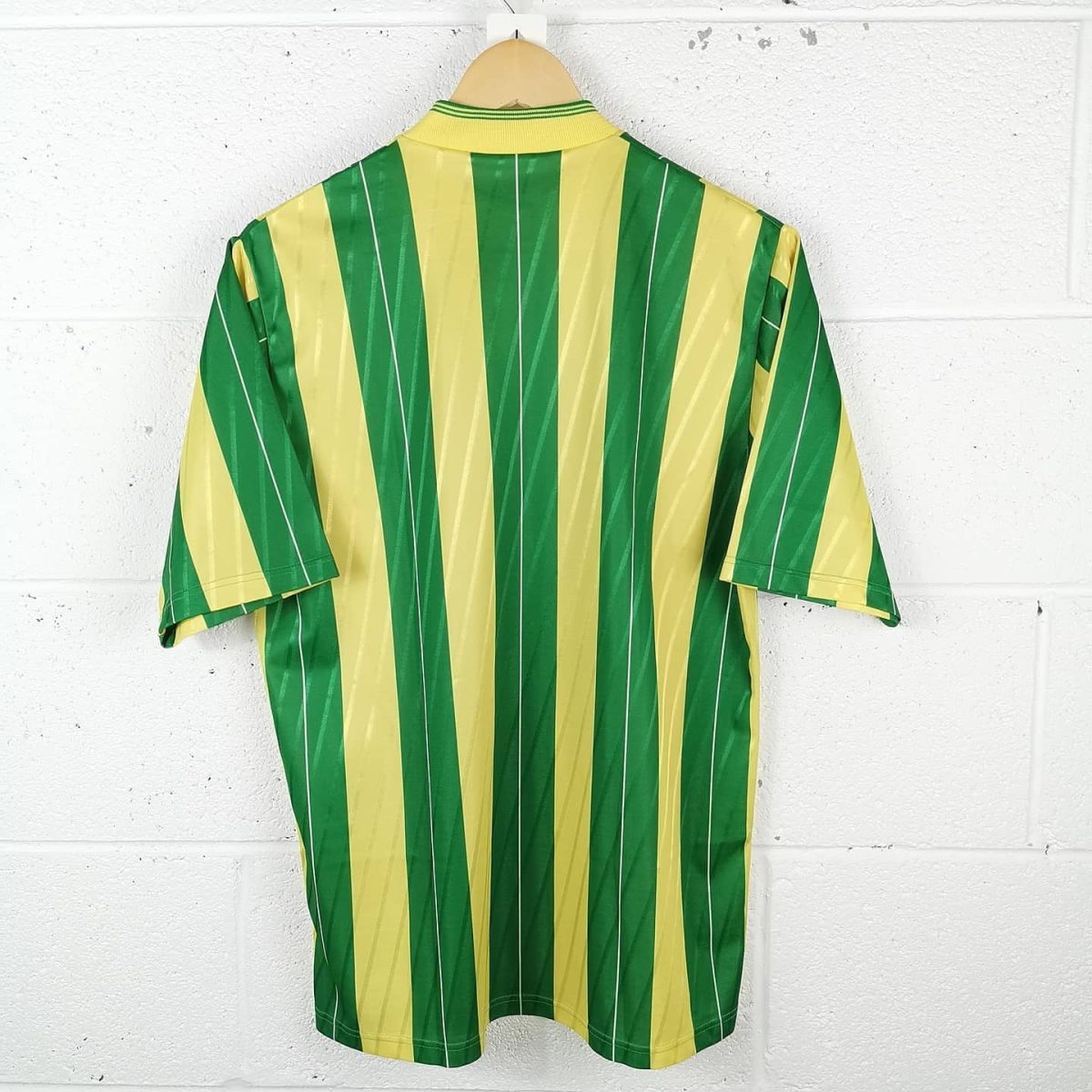 Green and yellow vertical striped shirt hanging on a wooden hanger against a white brick wall, product photo.