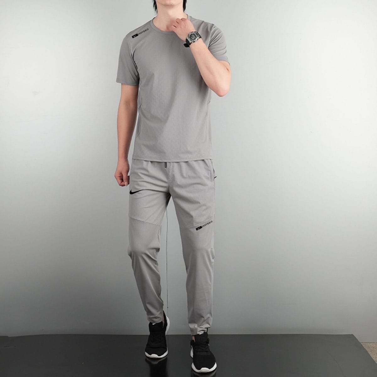 Person standing in a gray athletic outfit with Nike logos on the chest and pants, wearing black sneakers on a dark platform.