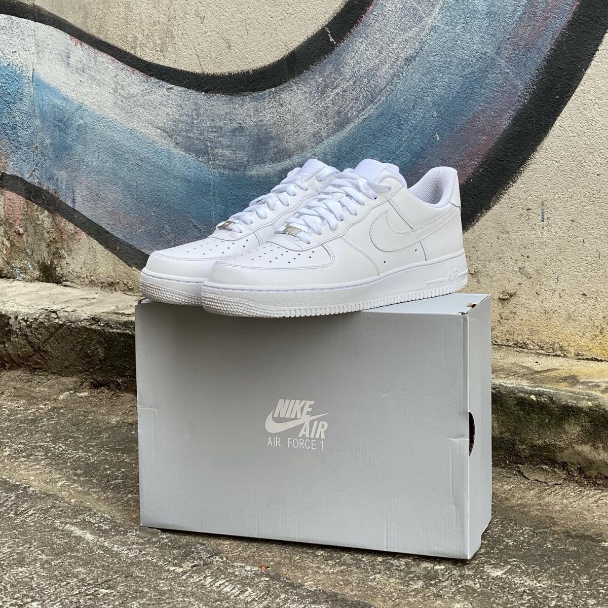 White Nike Air Force 1 sneakers resting on a gray shoebox outdoors against a graffiti wall backdrop on concrete ground.