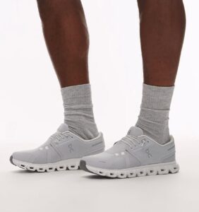 136456e0-39a2-4f47-97f7-39e926b341e0 | UAE SPORT STORE Light gray running shoes with thick white soles worn with gray crew socks, standing on a white background.
