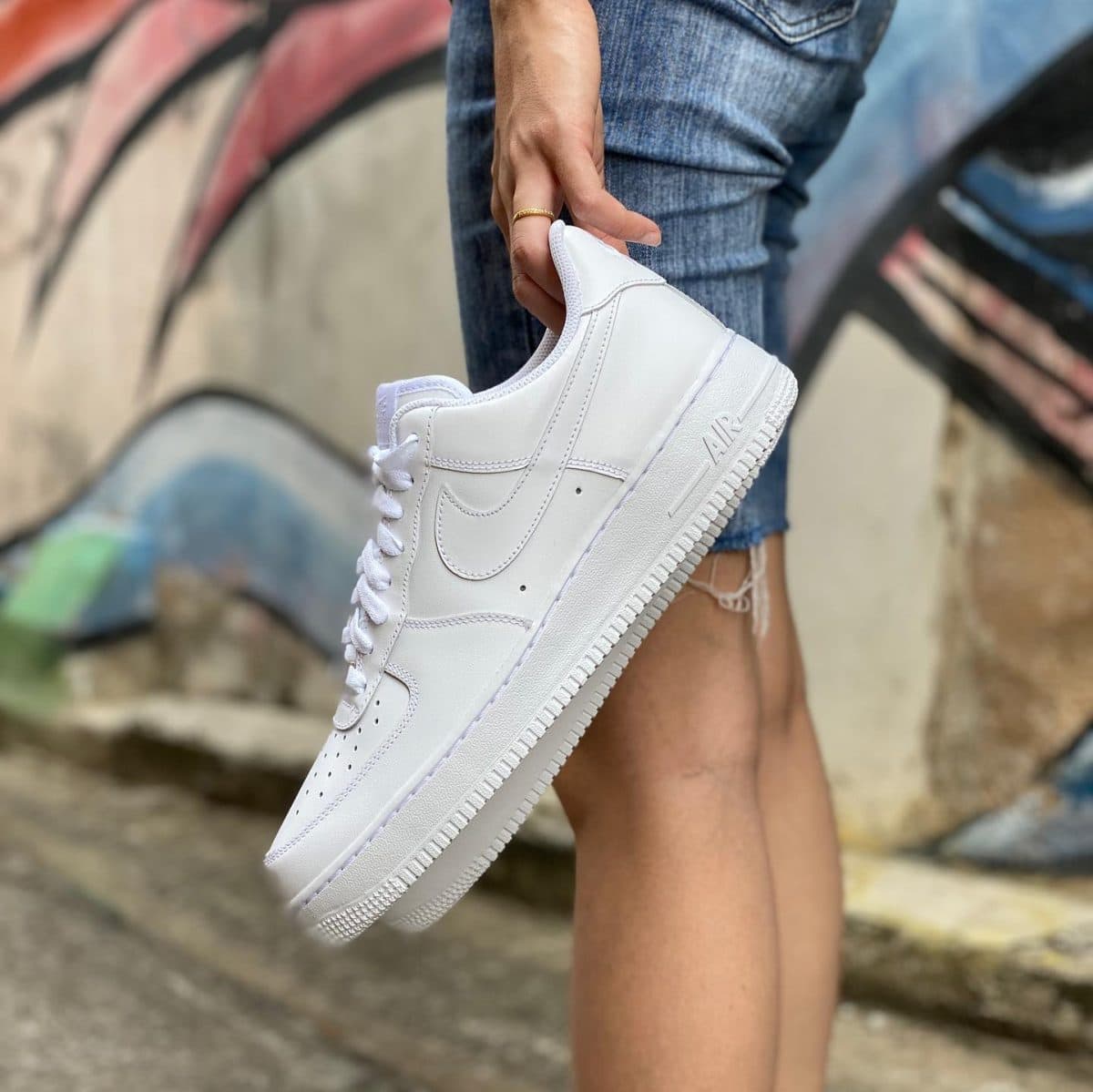 Close-up of a white Nike Air Force 1 sneaker being held by a hand in front of a graffiti-covered wall.