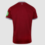 Back view of a red polo shirt with white-and-navy striped collar and sleeve cuffs, small crossed-tools logo at the neck, and a green circular patch on the left sleeve