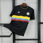 Black short-sleeve sports jersey with a yellow-blue-red horizontal stripe across the chest, Adidas logo on the left and a soccer-ball crest on the right, hanging on a wooden hanger against a metal grid backdrop.