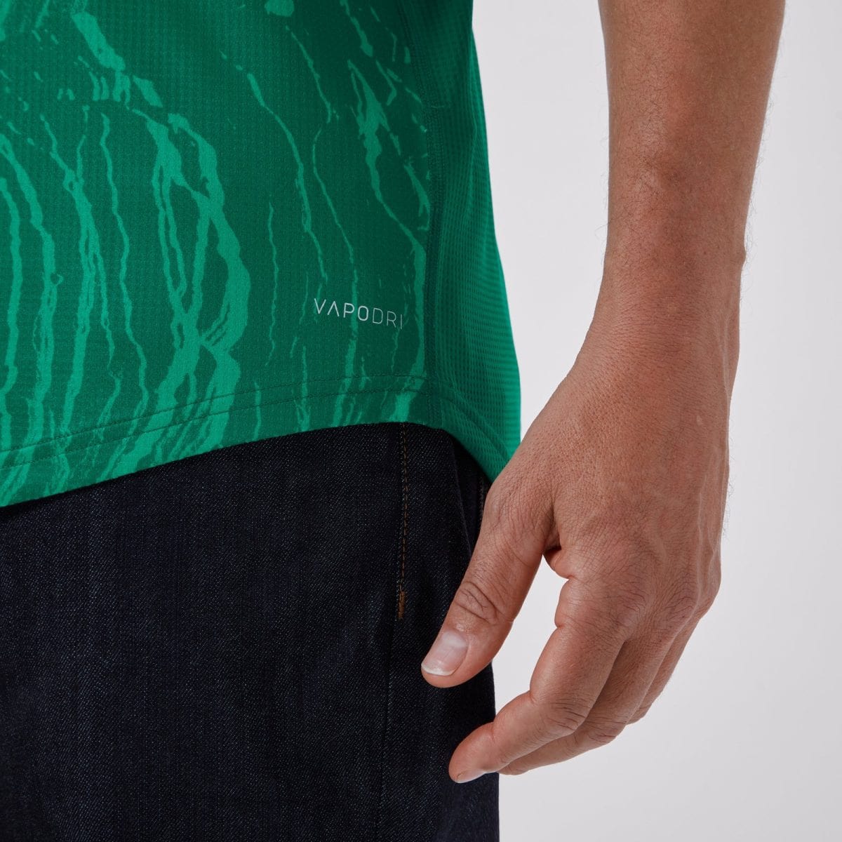 Close-up of a green patterned athletic shirt with the VAPODRI logo near the bottom hem, worn over dark jeans by a person.