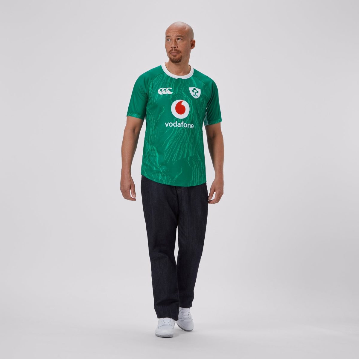 Man posing in a green rugby jersey with a white collar and Vodafone logo, standing against a light gray studio backdrop.