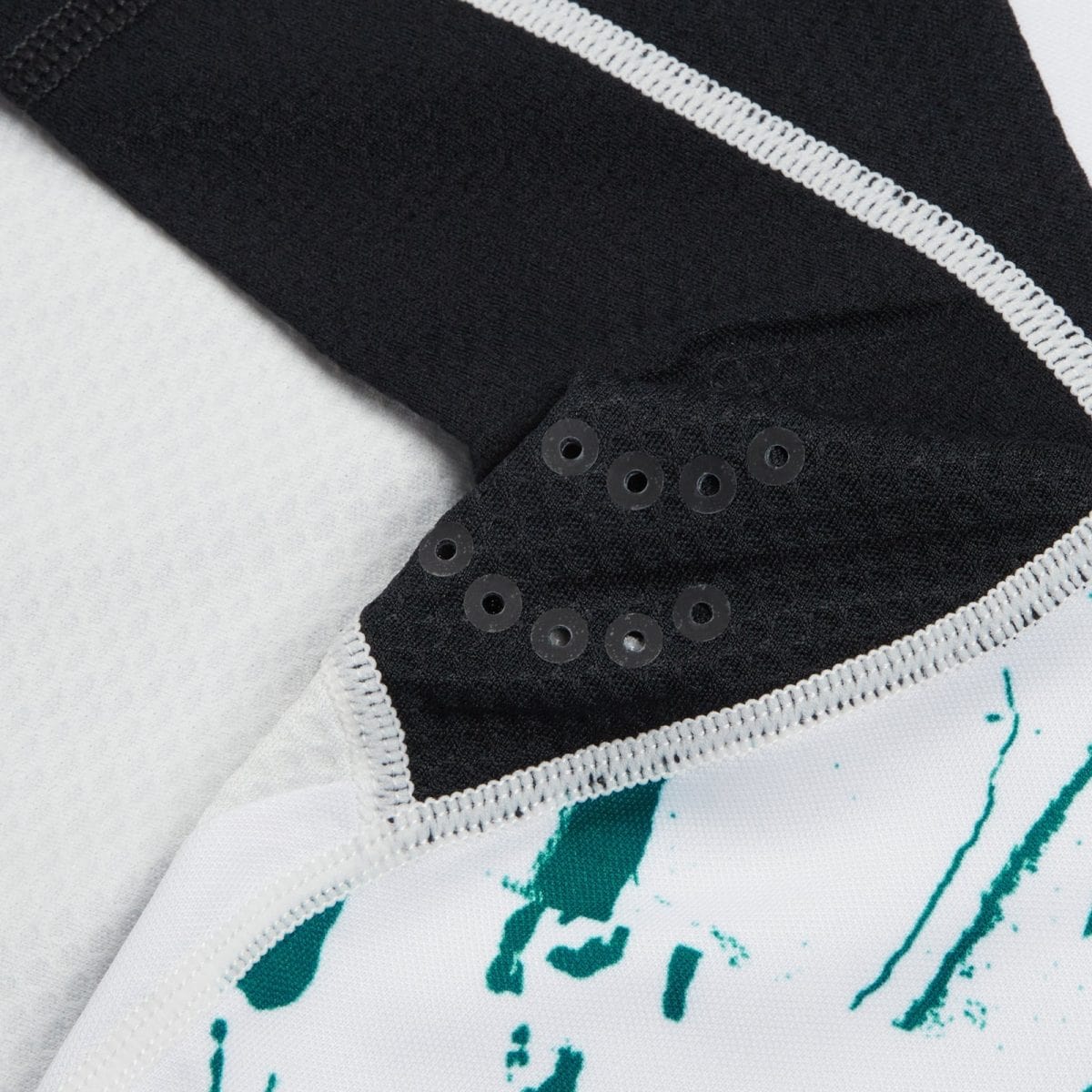 Close-up of a black sleeve with a row of metal eyelets along the cuff and a white stitched seam, resting on light fabric with teal paint marks.