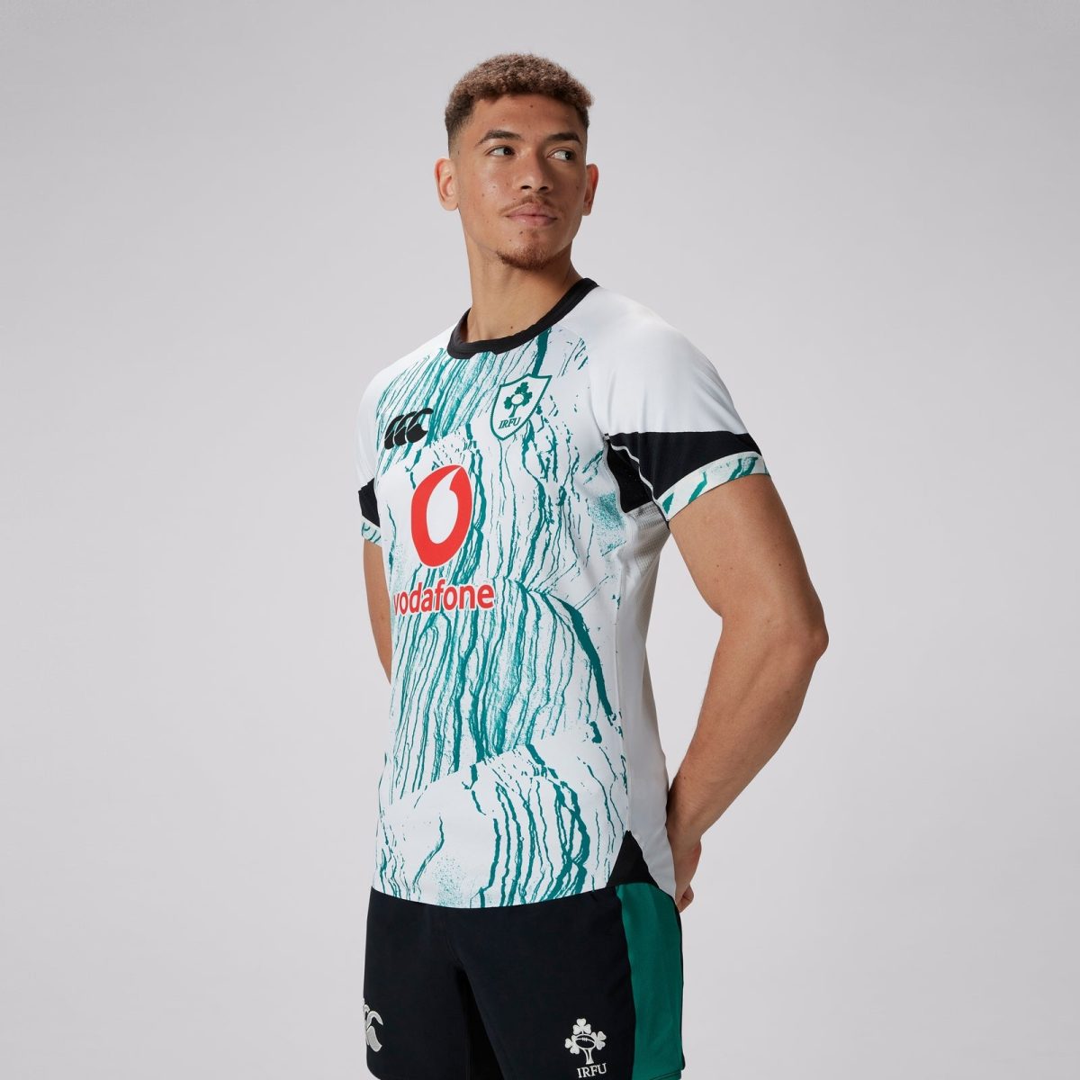Man wearing a white rugby jersey with teal splash pattern, Vodafone logo, and IRFU crest, looking to the left in a studio shot.