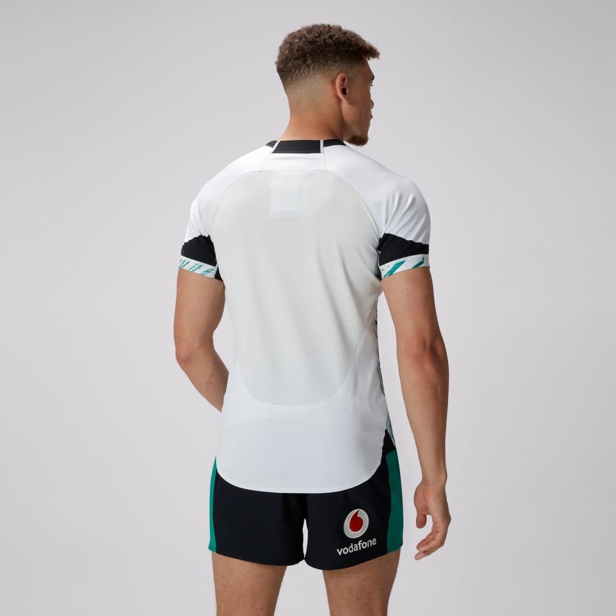 Man seen from behind wearing a white mesh-back athletic shirt and dark shorts with a Vodafone logo on the left thigh, neutral backdrop