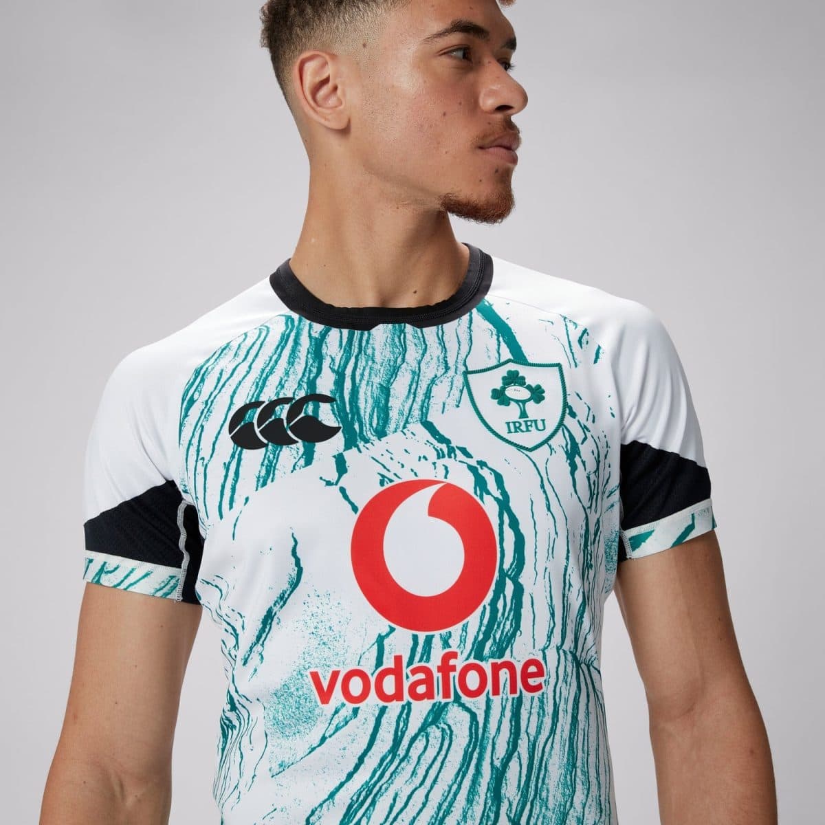 Man wearing a white teal-patterned rugby jersey with IRFU crest, Vodafone logo, and CCC emblem, looking to the right.
