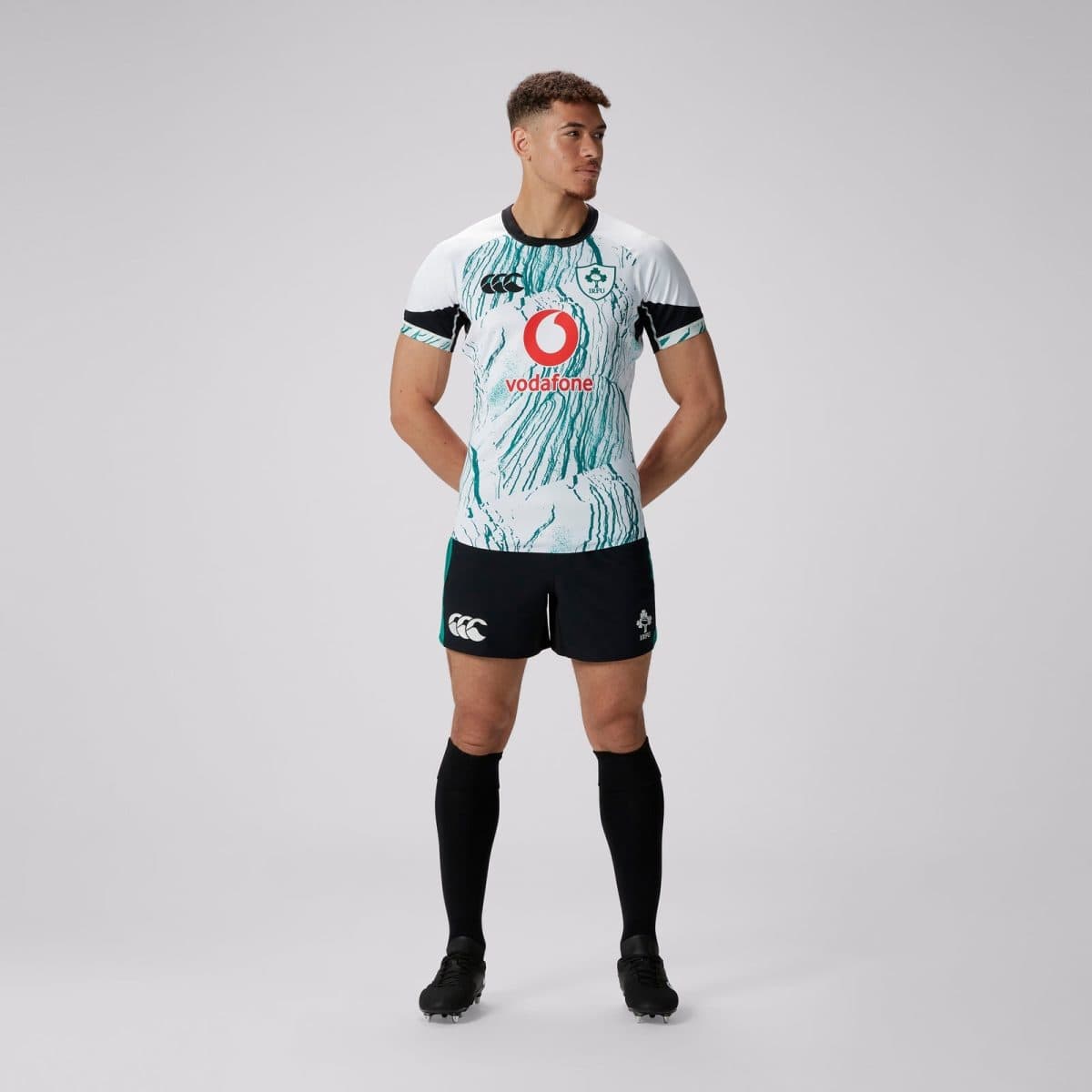 Rugby player wearing a white teal-pattern jersey with Vodafone logo and IRFU crest, black shorts, and black socks, facing slightly to the right with hands behind back.
