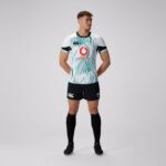 Rugby player wearing a white teal-pattern jersey with Vodafone logo and IRFU crest, black shorts, and black socks, facing slightly to the right with hands behind back.