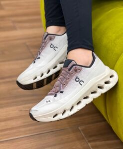14864e75-0616-4c7b-a4bc-8a9593a88a44 | UAE SPORT STORE Pair of white and blush athletic sneakers with pink laces and a chunky cut-out midsole, worn by someone in black leggings on a wooden floor beside a yellow chair.