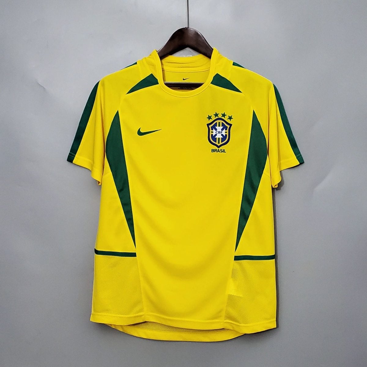 Yellow Brazil national football team jersey on a hanger, with green accents and team crest visible on the chest against a plain gray wall.