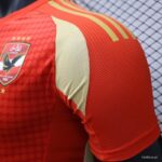 Close-up of a red sports jersey shoulder with a cream panel, featuring a shield crest on the left chest and yellow stripes across the back.