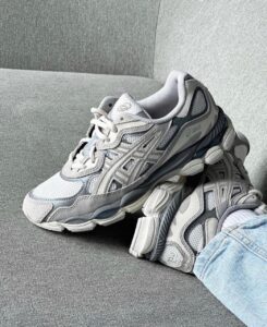 Grey and white chunky ASICS sneakers resting on a gray couch, with a jeans-clad leg nearby.