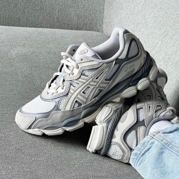 Grey and white chunky ASICS sneakers resting on a gray couch, with a jeans-clad leg nearby.