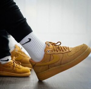 Close-up of a tan Nike sneaker on a foot wearing a white sock, with another sneaker in the background on a wooden floor.