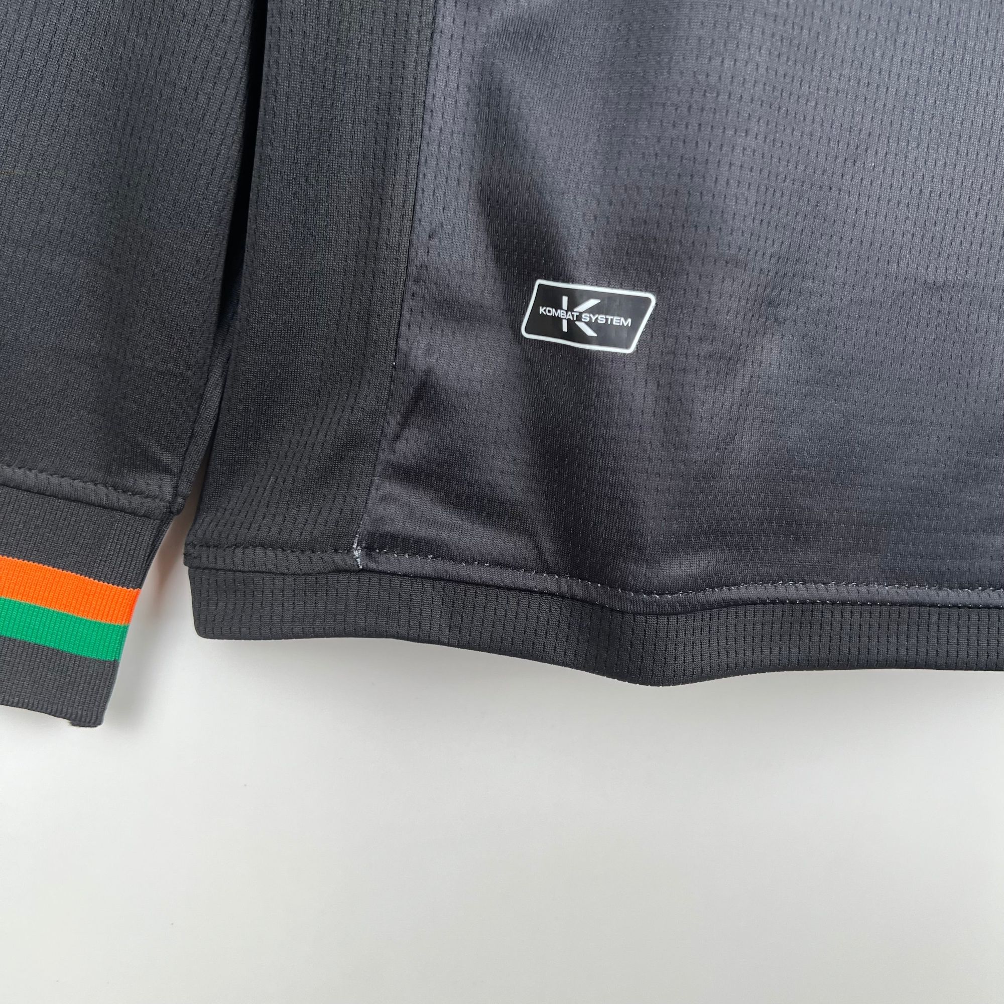 1528e7f7 | UAE SPORT STORE لقطة مقرّبة لقماش داكن مُحاك بملمس مع رقعة سوداء صغيرة مكتوب عليها "KOMBAT SYSTEM" قرب حافة سوار مخطط بالبرتقالي والأخضر.