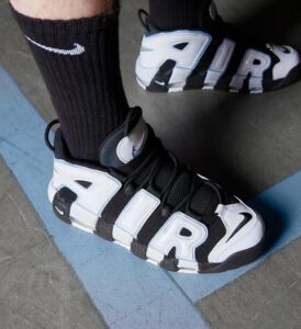 Close-up of black-and-white sneakers with large AIR lettering, worn with black socks on a gray floor with a blue curved line