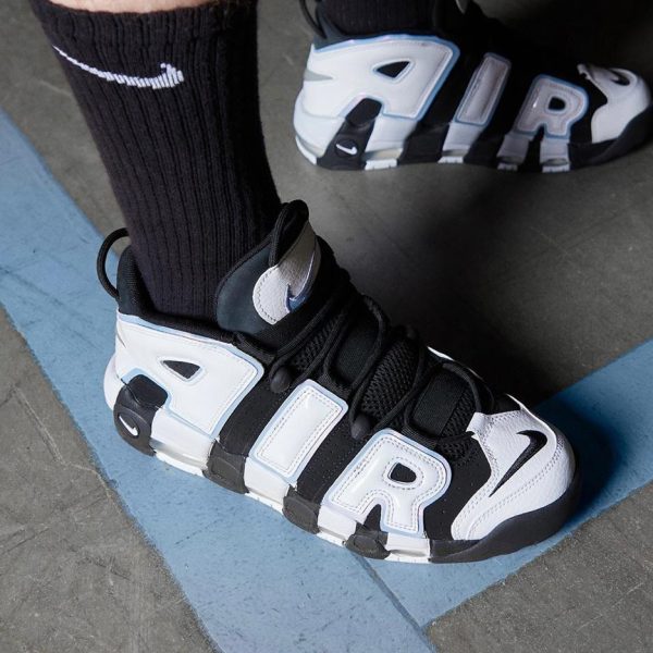 Close-up of black-and-white sneakers with large AIR lettering, worn with black socks on a gray floor with a blue curved line