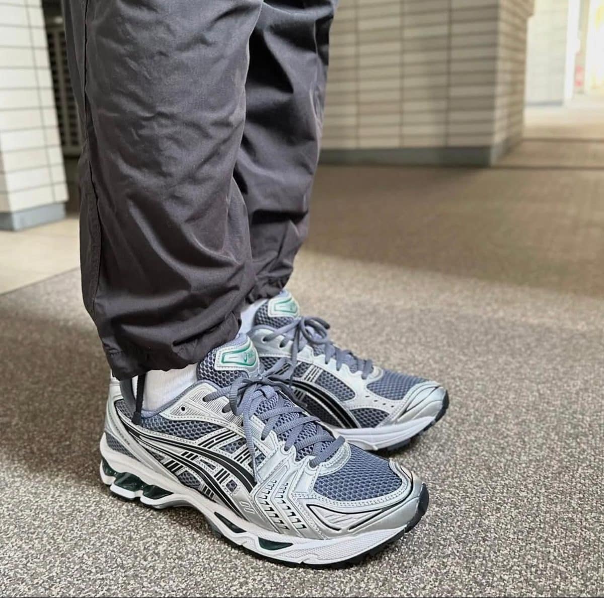 Close-up of gray and teal running shoes with mesh uppers, worn with black jogger pants in a tiled indoor hallway.]