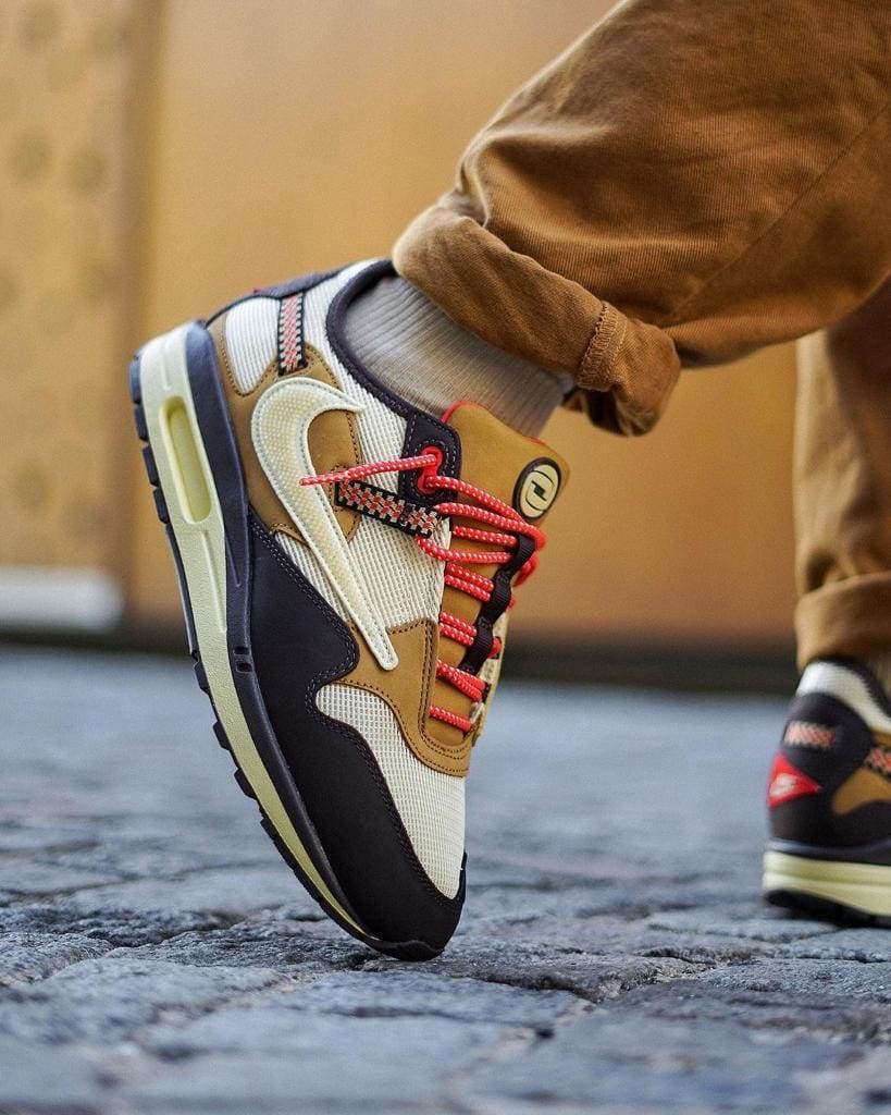 15cfbea7-2813-4aef-aef0-b21c3245b751_6ffbff56-1921-4547-a1f2-fcb6c8a4ec3d | UAE SPORT STORE Close-up of a multi-color sneaker with red laces resting on a cobblestone street, leg bent.