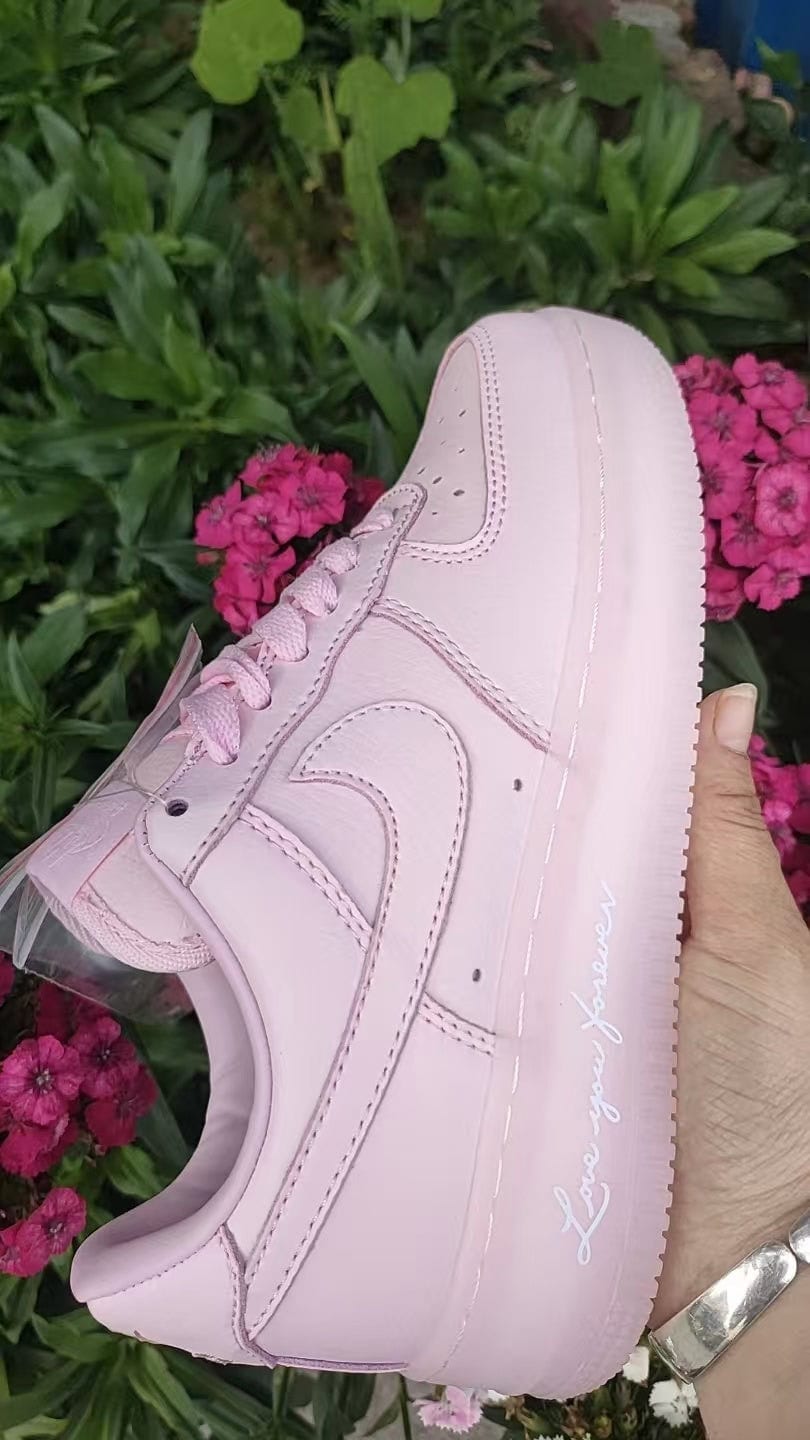 15db34c0 | UAE SPORT STORE Pastel pink low-top sneaker held up outdoors, featuring a white stitched Nike swoosh and cursive text along the midsole against a backdrop of pink flowers and green foliage.
